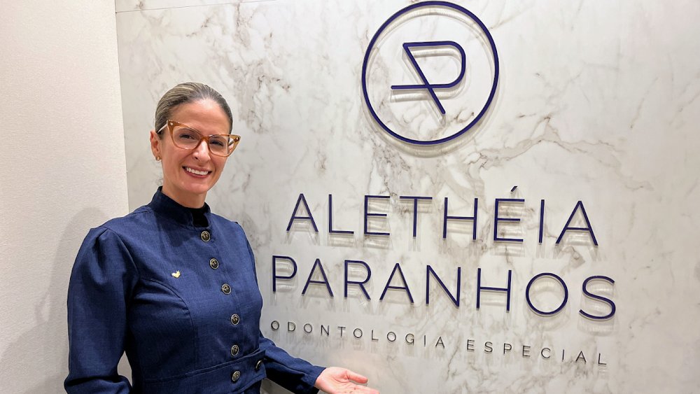 Dra. Alethéia Paranhos de uniforme azul-marinho ao lado da marca Alethéia Paranhos Odontologia Especial, instalada na parede marmorizada do consultório